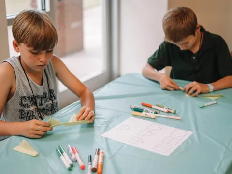 a couple of boys sitting at a table with a blue table with pencils and paper