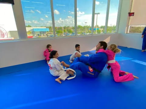 a group of children sitting on a blue mat in a room with large windows