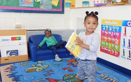 a young girl holding a book and a baby in a chair