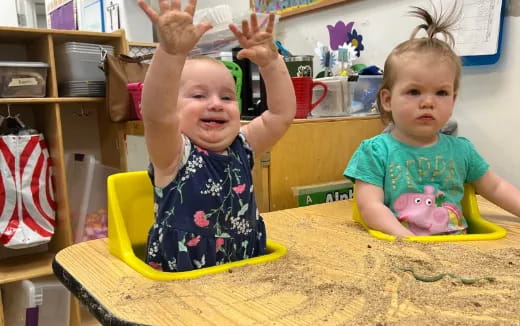 a couple of young children sitting at a table with their hands up
