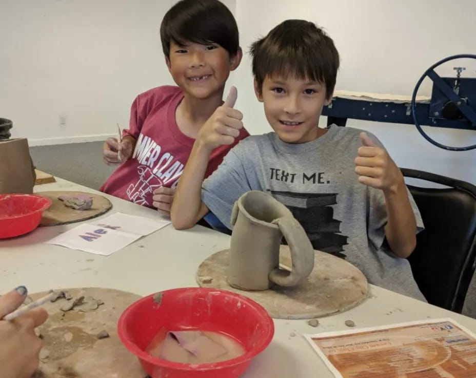 a couple of kids sitting at a table with a clay pot and a bowl of food