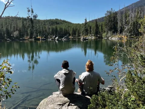 a couple of people sitting on a rock looking at a lake with trees and mountains in the background