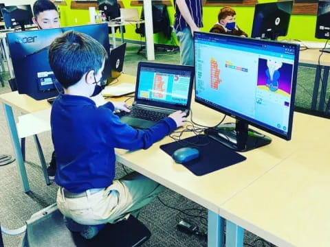 a boy sitting at a desk with a computer and a laptop