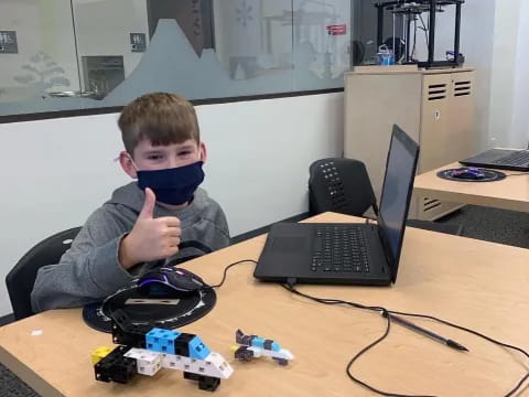 a boy with a mask on his face sitting at a desk with a laptop and a computer