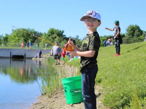 a boy holding a bucket and a fishing pole by a pond