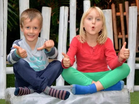 a boy and girl sitting on a bench and pointing at the camera