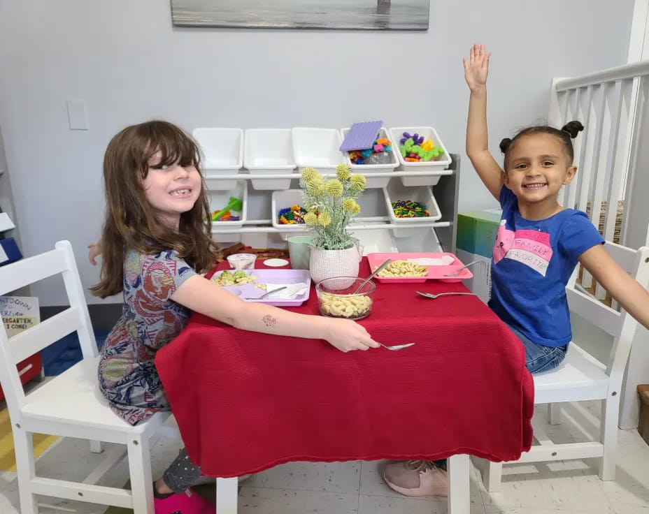 a couple of children sitting at a table with a red tablecloth and a white chair with a