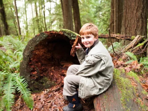 a boy squatting next to a large rock in the woods