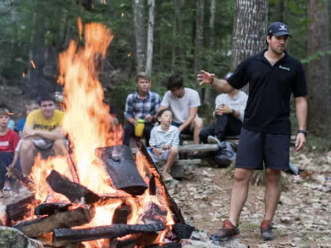 a person standing next to a campfire with people sitting around
