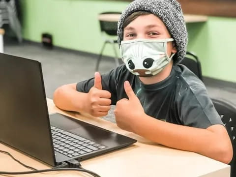 a person wearing a mask and sitting at a table with a laptop