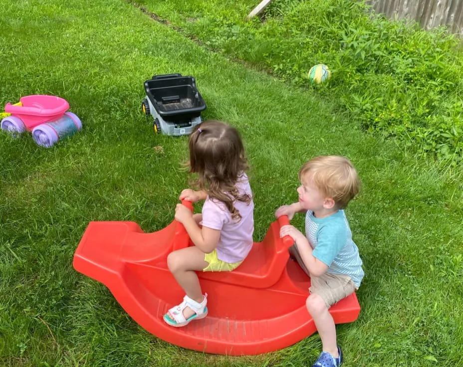 two children sitting on a red plastic toy in a grassy area