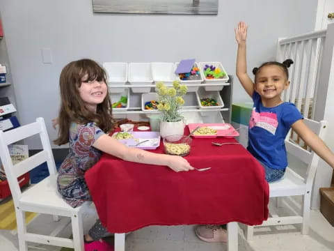 a couple of children sitting at a table with a red tablecloth and a white chair with a