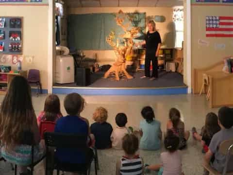 a group of children sitting in a room with a person standing in front of a stage