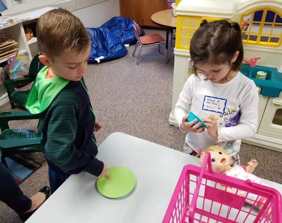 a couple of kids sit at a table with a doll