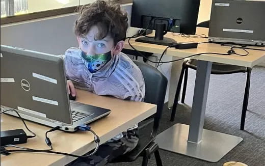 a boy with a face mask sitting at a desk with a laptop