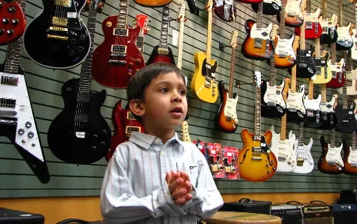 a boy sitting in front of a wall of guitars