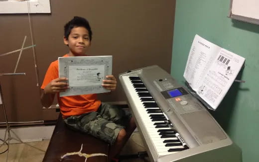 a boy holding a paper and sitting next to a piano