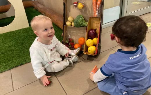 a couple of babies sitting on the floor by a fruit stand