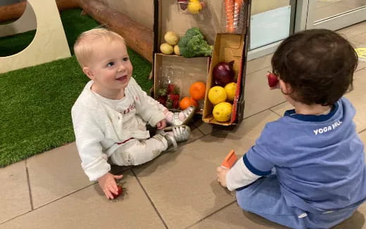 a couple of babies sitting on the floor by a fruit stand