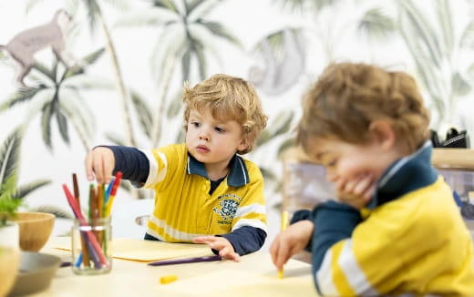 a couple of young boys sitting at a table with pencils and a plant