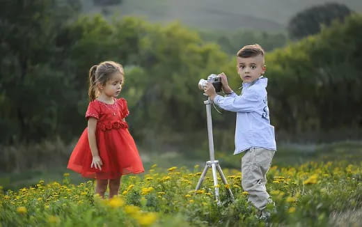 a boy and girl standing in a field of flowers and looking at a camera